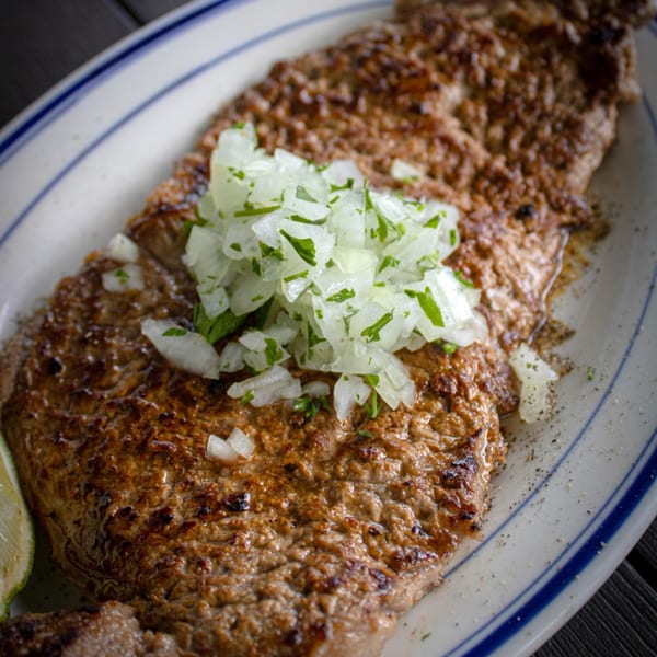Grilled steak topped with diced white onions and parsley, served on a white plate with blue stripes.