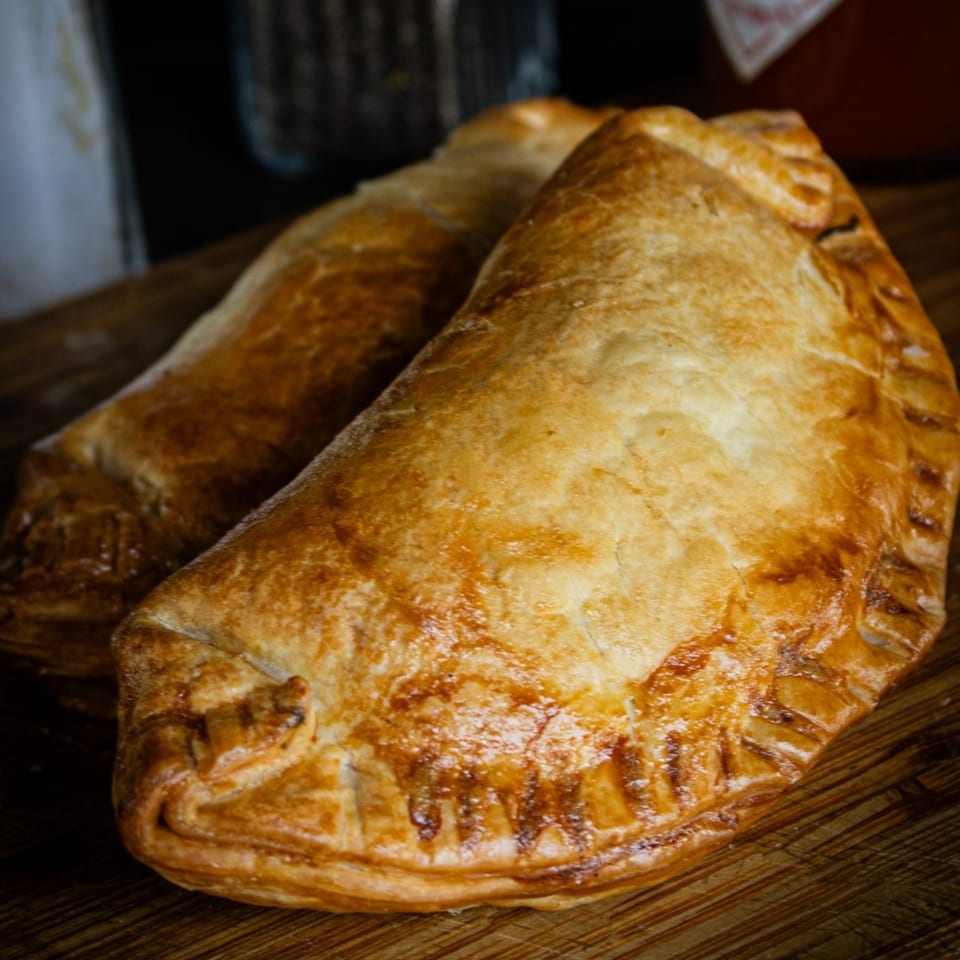 Two golden-brown, flaky pastries, possibly empanadas or pasties, resting on a wooden surface.