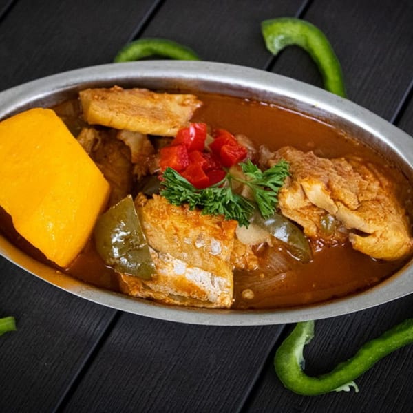A metal dish filled with chicken stew, green peppers, and a yellow plantain on a dark wooden table.
