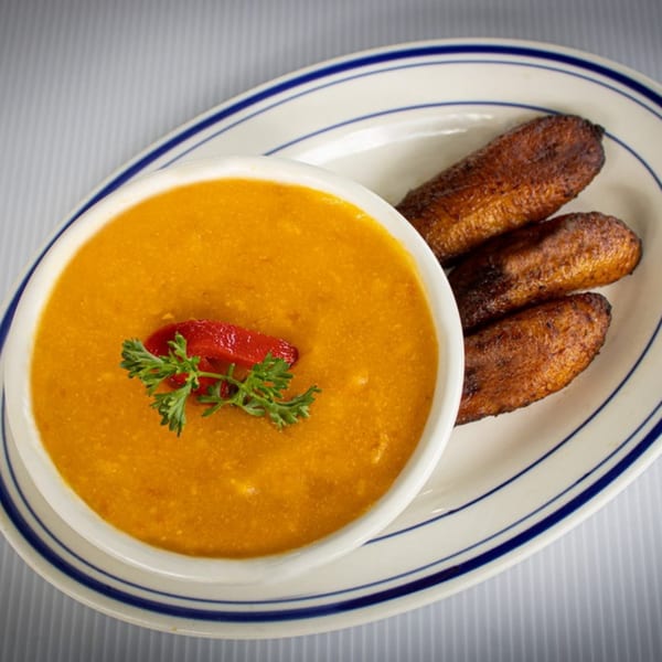 A bowl of orange soup with red pepper and parsley, served with three fried plantains.