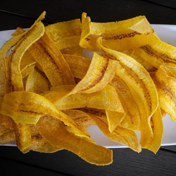 Close-up of golden-brown plantain chips on a white rectangular plate, against a dark background.