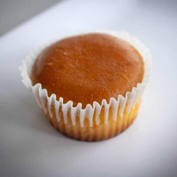 A single, golden-brown cupcake in a white paper liner sits on a white surface.