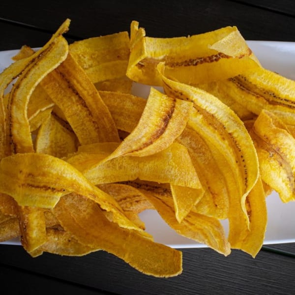 A white plate piled high with golden-yellow, crispy plantain chips on a dark wooden surface.