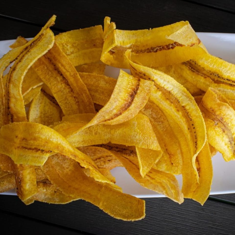 A white plate piled high with golden-yellow, crispy plantain chips on a dark wooden surface.