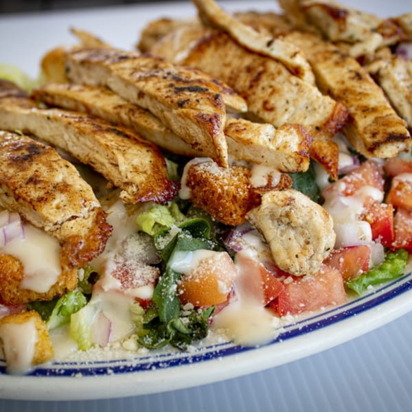 Grilled chicken Caesar salad with croutons, tomatoes, red onion, and dressing on a white plate.