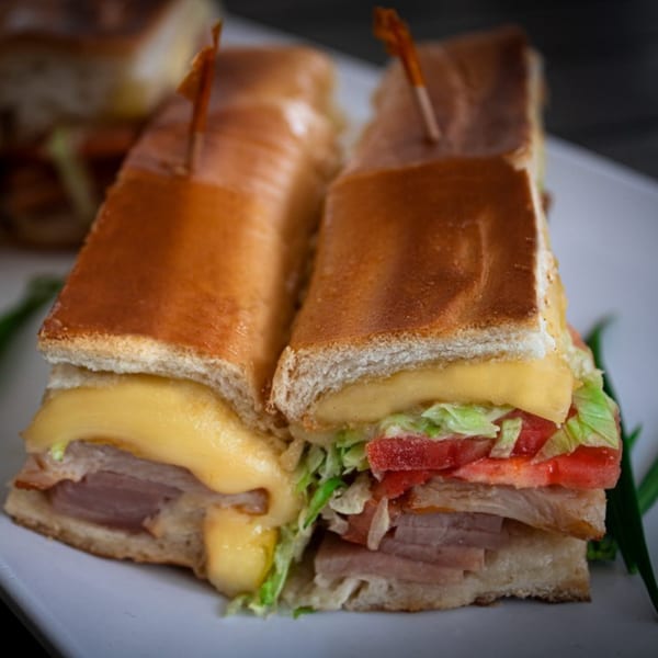 Two halves of a Cuban sandwich with melted cheese, ham, lettuce, and tomato on a white plate.