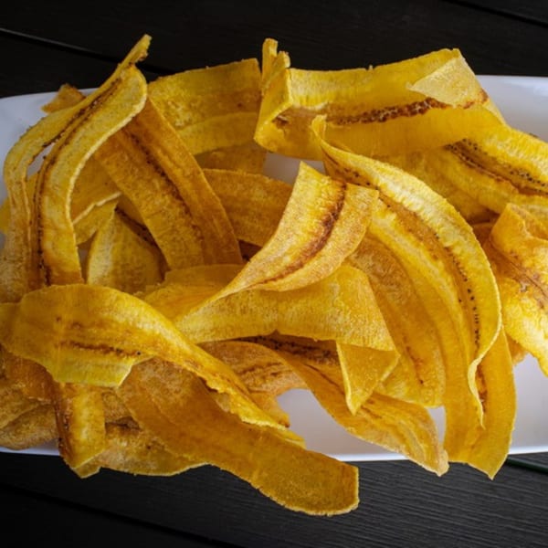 A white plate filled with golden-brown, crispy plantain chips on a dark wooden table.