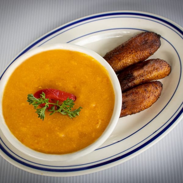 A bowl of orange soup garnished with parsley and red pepper, served with fried plantains.