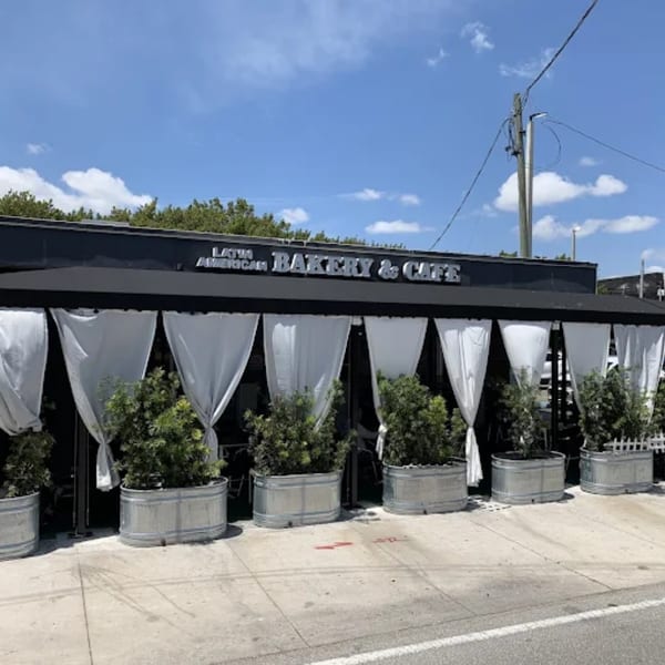 Exterior of Latin American Bakery & Cafe, with a black awning, white curtains, and potted plants.
