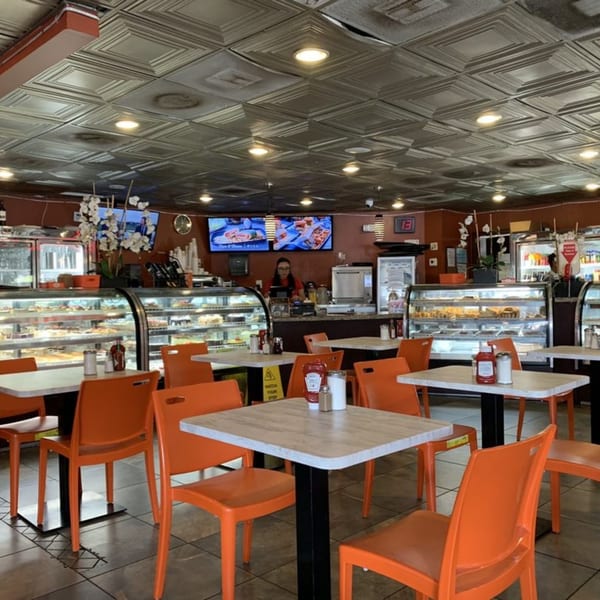 A brightly lit diner interior with orange chairs, white tables, and display cases filled with pastries.