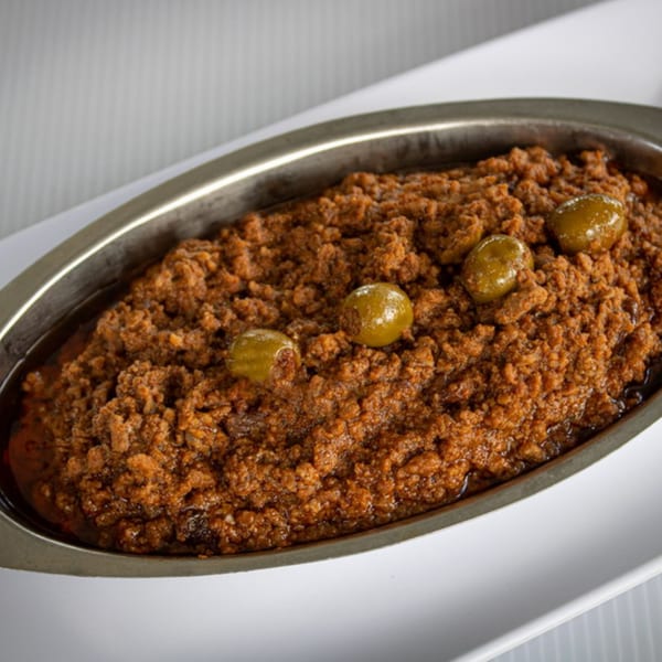 Savory brown ground meat dish with green olives in a silver serving dish on a white plate.